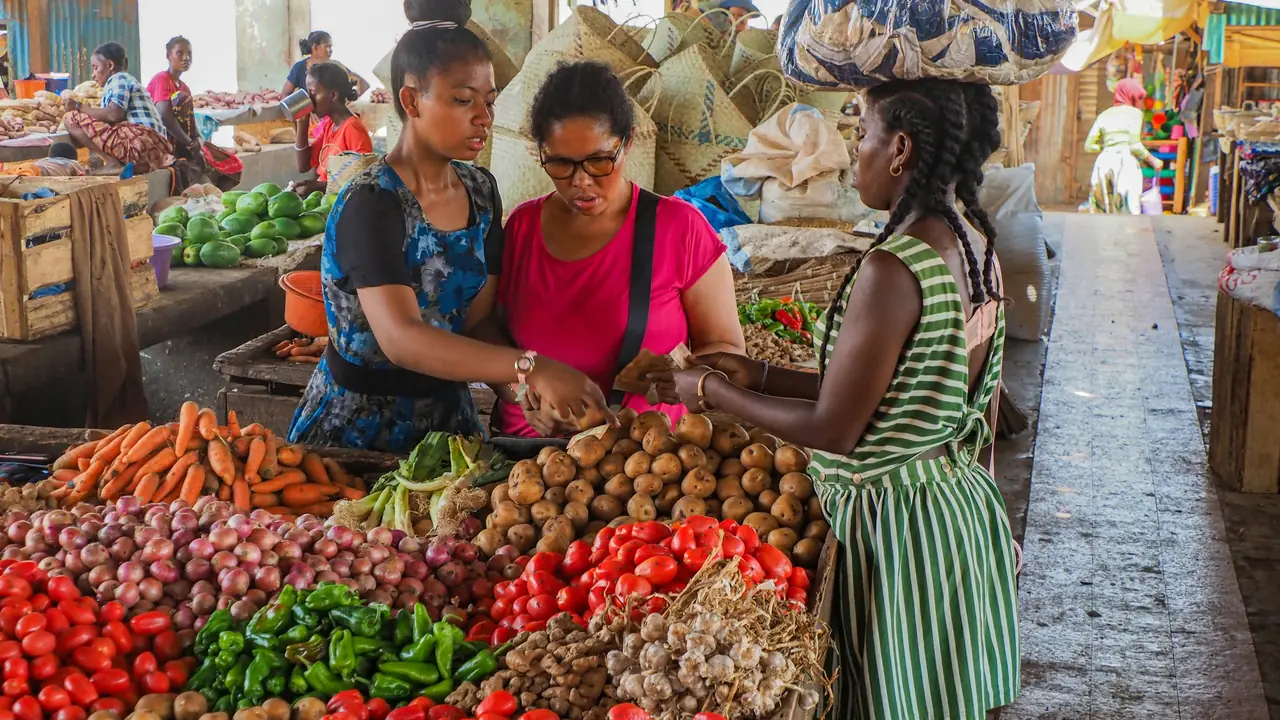 Trois clientes choisissent des tomates, des carottes et des oignons frais sur un étal de marché coloré, en plein jour.